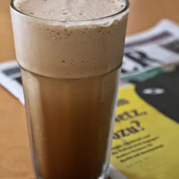 A tall glass of iced coffee with a thick foamy top.