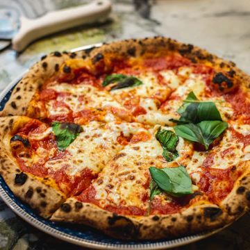 A round pizza with a puffy, charred crust, tomato sauce, melted mozzarella and fresh basil leaves.