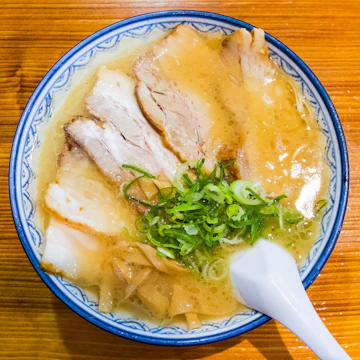 A bowl of cloudy pork broth with noodles, sliced pork belly, spring onion and bamboo shoots, with a spoon.